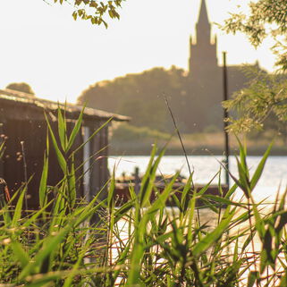 Schilf im Vordergrund, dahinter Bootsschuppen, Wasser und Kirche